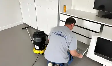 A cleaner from Housecleaners.nl kneels to clean the bottom shelf of a white wardrobe, with a large industrial vacuum standing next to him